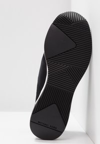 Semelle d'une basket noire avec une semelle en caoutchouc texturée et le logo "Michael Kors" près du talon, sur fond blanc.