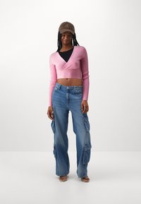 Rosa gerippter, verkürzter Pullover mit langen Ärmeln, kombiniert mit hellblauen Cargo-Jeans mit mehreren Taschen. Das Outfit wird mit einer braunen Mütze abgerundet.