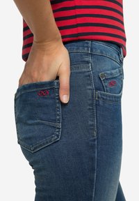 Tummansiniset keskikorkeat farkut räätälöidyllä leikkauksella, joissa on punaiset kirjailut takataskuissa ja näkyvät tikit.