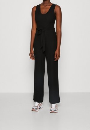 Czarny prążkowany kombinezon z dekoltem w serek, bez rękawów i z wiązanym paskiem w talii, w połączeniu z kolorowymi butami sportowymi.