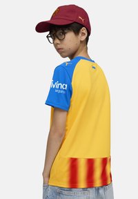 Kille som bär glasögon, en vinröd keps med logotyp och en gul-blå sporttröja med röda ränder på nedre delen av ryggen, som vänder sig åt sidan.