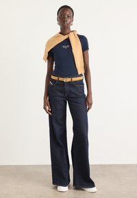 Marine T-skjorte med tekst, gul strikkegenser drapert, vide mørke jeans, gul belte, hvite sneakers, glatte teksturer, uformelt antrekk.