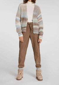 Sweter z dzianiny w kolorze beżowym, brązowym i niebieskim z guzikami, w połączeniu z brązowymi spodniami z ekoskóry oraz tanowymi botkami za kostkę z pętelkami i teksturowanymi skarpetkami.