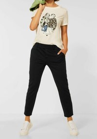 Beige grafisk t-shirt med en illustration av två kvinnor, i kombination med svarta smala byxor och vita sneakers på en neutral bakgrund.