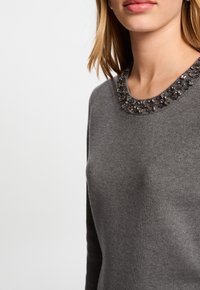 Grauer Pullover mit einem Rundhalsausschnitt, der mit einem Perlenbesatz verziert ist. Der Stoff wirkt weich und hat eine feine Textur.