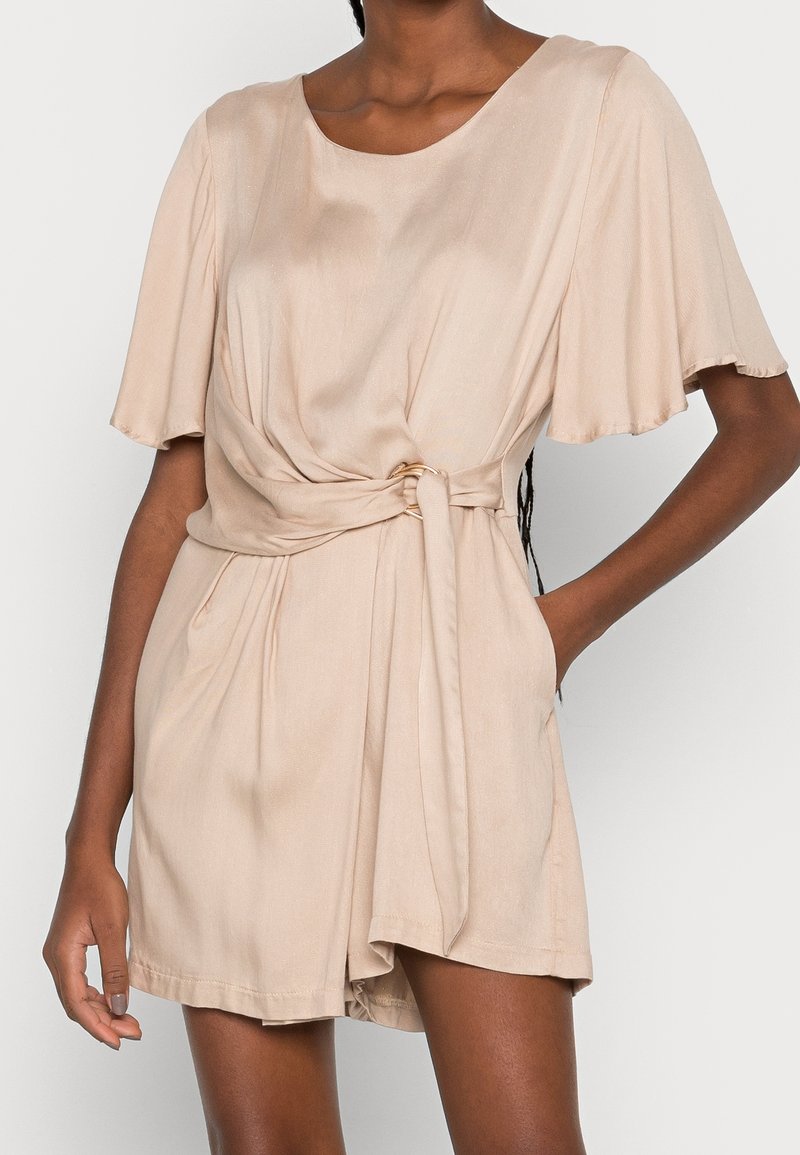 Femme portant une robe beige à manches courtes avec une ceinture nouée à la taille et une jupe légèrement évasée sur un fond clair uni.