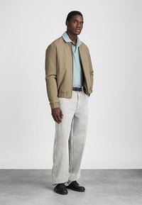 Μπεζ bomber τζάκετ πάνω από ένα ανοιχτό μπλε πόλο, σε συνδυασμό με ανοιχτό γκρι παντελόνι wide-leg και μαύρα παπούτσια. Απλός, σύγχρονος σχεδιασμός χωρίς ορατά μοτίβα.