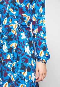Blaues Blumenwickelkleid mit langen Ärmeln, das mit weißen, gelben und dunkelvioletten Blütenmustern verziert ist. Der gebundene Taillendetail betont die Figur.