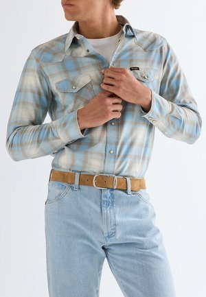 Mann knöpft ein hellblau-beige kariertes Langarmhemd über ein weißes T-Shirt, trägt hellblaue Jeans und einen braunen Wildledergürtel mit silberner Schnalle.