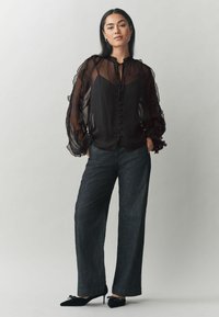 Blusa nera trasparente con maniche a sbuffo e frontale con bottoni, abbinata a pantaloni in denim scuri a gamba larga e décolleté neri a punta.
