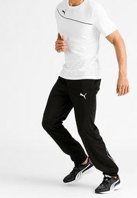 Vit idrottströja med svarta accenter och logotyp, kombinerad med svarta byxor med sidostråk och svarta sneakers med vita detaljer.