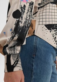 Langärmlige Bluse aus cremefarbenem Stoff mit abstrakten schwarzen und bunten Spritzmustern. Mit abgerundetem Saum und gestreiften Bündchen, kombiniert mit blauen Jeans.