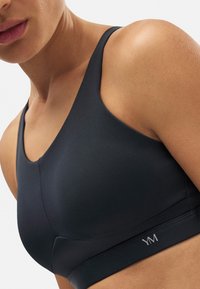 Sujetador deportivo negro con una textura suave y mate, que presenta tirantes anchos, un escote curvado y un pequeño logo en el dobladillo.