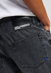 Crne traperice od denima s teksturiranim završetkom, s crnom kožnom etiketom na pojasu koja prikazuje "KARL LAGERFELD JEANS" u bijelom tekstu.