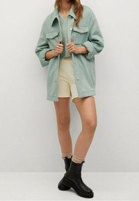 Giacca oversize verde menta, top in maglia, pantaloni pantaloni beige, stivali neri alla caviglia con calze beige, con dettagli a bottoni e vestibilità comoda.