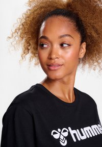 Schwarzer Sweatshirt aus Baumwolle mit Rundhals und einem weißen Logodesign auf der Brust. Modell mit lockigem Haar, das sich leicht gedreht hat.