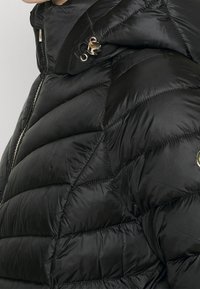 Schwarze Steppjacke mit hohem Kragen, Reißverschluss und glänzender Textur, mit gesteppten Polstern und einem dekorativen Metallakzent.