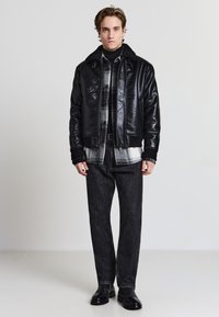 Schwarze Lederbomberjacke mit einem Kunstfellkragen, über einem karierten Hemd getragen, kombiniert mit schwarzen Jeans und schwarzen Stiefeln.