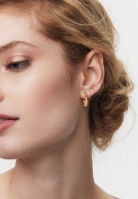 Złote kolczyki koła mają teksturowany wzór z gładkim wykończeniem. Kolczyki mają owalny kształt, co podkreśla elegancję noszącej je osoby.