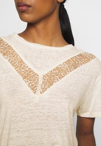 Mujer vestida con una blusa de manga corta color crema con un detalle de encaje en forma de V en el pecho y los hombros, con el pelo recogido en una coleta.
