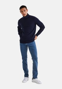 Pull col roulé bleu marine en matériau doux, associé à un jean en denim bleu et des baskets blanches. Le design est ajusté, avec des poignets côtelés.