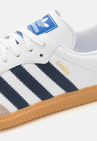 Witte Adidas Samba sneaker met navy drie strepen, gum rubberen zool en blauw Adidas logo op de tong.