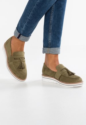Loafers suede hijau zaitun dengan detail tassel, dilengkapi dengan sol karet putih dan aksen jahitan cokelat, dipadukan dengan celana jeans denim gulung.