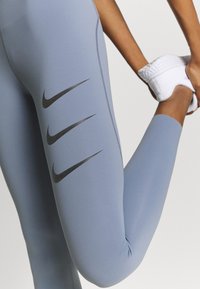 Leggings de sport bleues avec une texture lisse, un matériau élastique et quatre logos Nike Swoosh noirs sur la jambe inférieure.