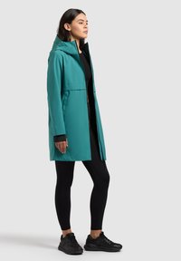 Frau steht linksblickend, trägt einen türkisfarbenen Regenmantel mit Kapuze über schwarzen Leggings und schwarzen Sneakers vor hellgrauem Hintergrund.