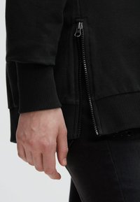 Black sweatshirt featuring a side zipper, ribbed cuffs, and a smooth texture, with a close view of the hand near the zipper detail.