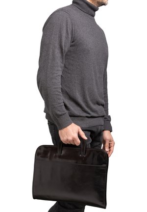 Man in een grijze coltrui die een zwarte leren aktetas bij het handvat vasthoudt, staand tegen een witte achtergrond.