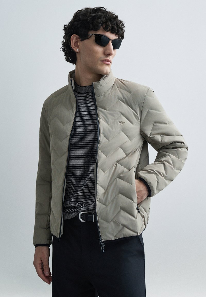 Bézs steppelt dzseki magas gallérral, cipzáras lezárással és fekete béléssel. Egy mintás fekete-fehér inggel és sötét nadrággal párosítva.