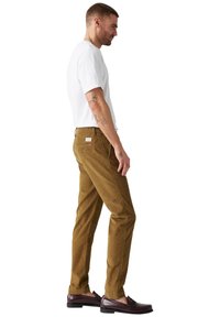 Pantaloni in corduroy marrone con vestibilità dritta, dotati di cinque tasche e un'etichetta con il marchio. Indossati con una t-shirt bianca semplice e mocassini.