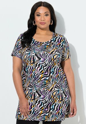 Kobieta z długimi ciemnymi włosami, nosząca kolczyki koła i kolorową bluzkę w paski zebry z krótkim rękawem, stojąca na tle gładkiej ściany.