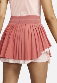 Primer plano de una persona que lleva una falda de tenis plisada rosa con pantalones cortos integrados y una cinturilla de malla, contra un fondo blanco.