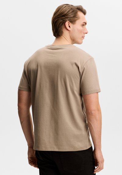 Uomo con capelli castano chiaro che indossa una maglietta beige a maniche corte, visto di spalle su uno sfondo bianco uniforme.