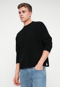 Suéter negro de canalé con cuello redondo y hombros caídos, combinado con jeans azul claro. La tela parece texturada y acogedora.