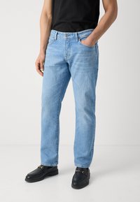 Jean en denim bleu clair avec une coupe décontractée, design à cinq poches, décoloration subtile et fermeture à bouton à la taille. Associé à des mocassins noirs.