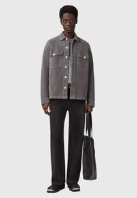 Grijze canvas jas met twee voorzakken en witte knopen, gecombineerd met zwarte jeans en een gestructureerde zwarte totebag. Zwarte schoenen maken de look af.