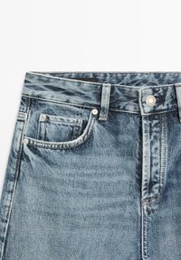 Blauwe denim jeans tailleband met een voorknoop, riemlussen en zichtbare stiksel op de voorzak op een witte achtergrond.