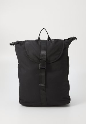Sac à dos en tissu noir doux avec rabat supérieur, fermeture à boucle réglable, côtés à cordon et une boucle de poignée sur fond clair uni.
