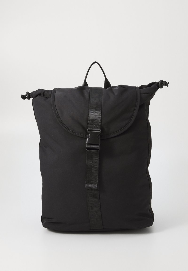 Sac à dos en tissu noir doux avec rabat supérieur, fermeture à boucle réglable, côtés à cordon et une boucle de poignée sur fond clair uni.