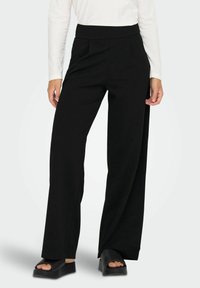 Sorte breddebukser lavet af blødt stof med høj talje, der har frynser foran og en glat tekstur. Matchet med sorte slidesandaler.