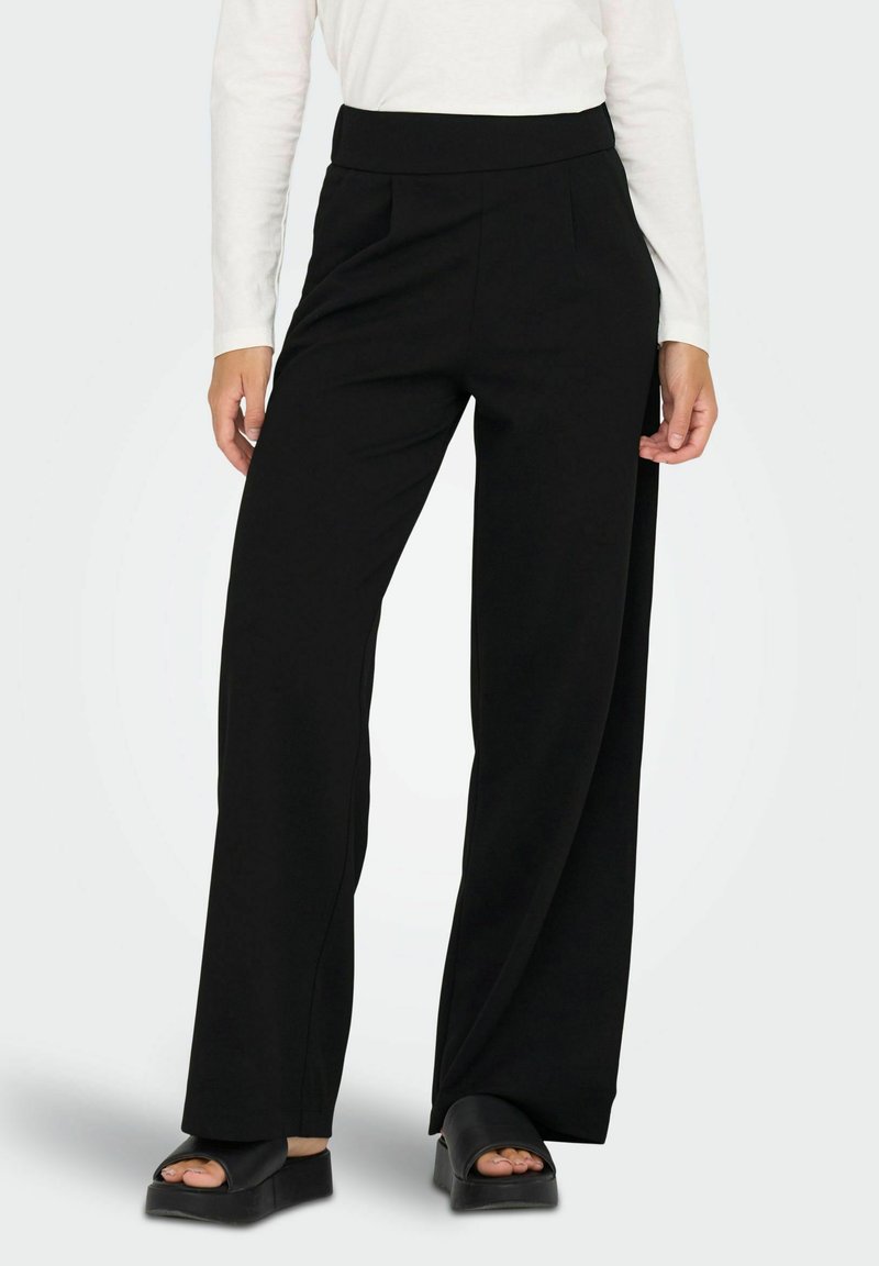 Sorte breddebukser lavet af blødt stof med høj talje, der har frynser foran og en glat tekstur. Matchet med sorte slidesandaler.