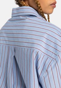 Light blue shirt with thin red and white vertical stripes. Features a collar and a loose fit design with soft textured fabric.
