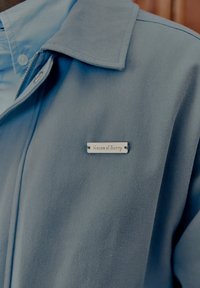 Close-up of a light blue jacket with a metal nameplate reading "House of Sunny" on the chest near the collar.