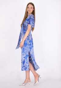 Robe paisley bleue à manches courtes, avec un design enroulé et une fente sur le côté, assortie à des chaussures à talons argentées. Tissu lisse et motifs vibrants.