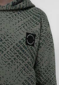 Felpa in tessuto verde oliva con motivo testuale ripetuto di colore verde scuro. Include un'etichetta con logo nera sul petto e design con cappuccio.