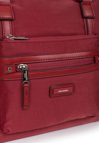 Borsa in nylon rossa con dettagli in pelle, dotata di una tasca con zip frontale, un'etichetta con la scritta "HEXAGONA" e spallacci regolabili per il trasporto.