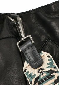 Zwarte leren tas met een zilveren haaksluiting. Heeft een geprint stoffen label in beige, zwart en groene tinten dat met een leren band is bevestigd.